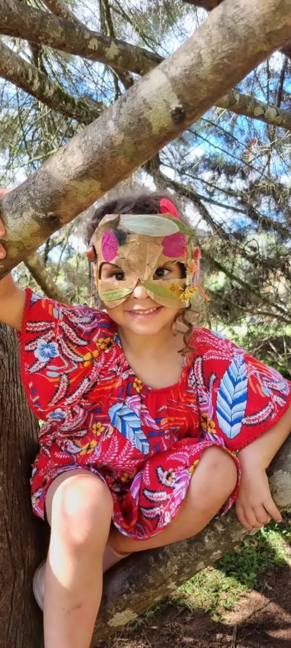 Menina sorridente de vestido vermelho e máscara de caixa com folhas subida no tronco de uma árvore