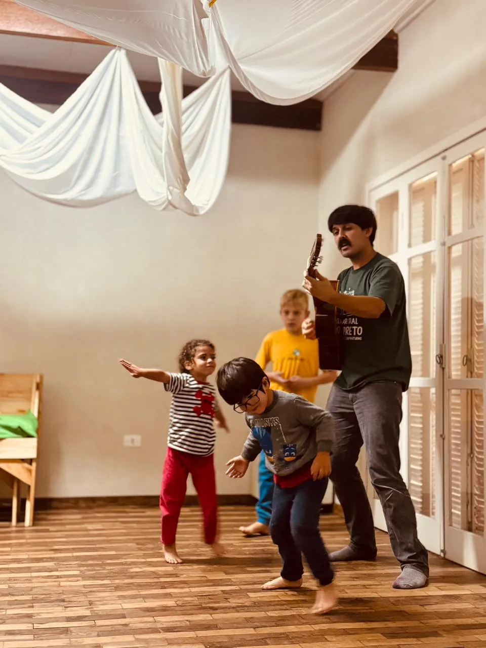 Homem tocando violão e cantando enquanto crianças pequenas dançam felizes pela sala