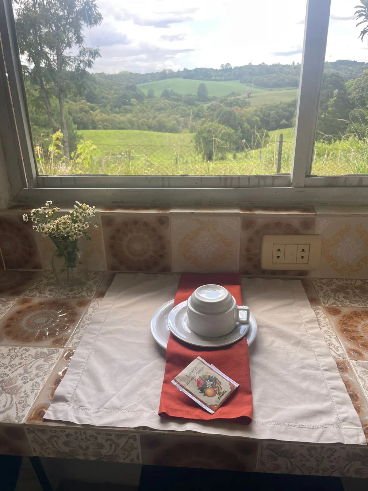 Xícara branca e um sachê de chá em bancada azulejada com vista relaxante da serra pela janela