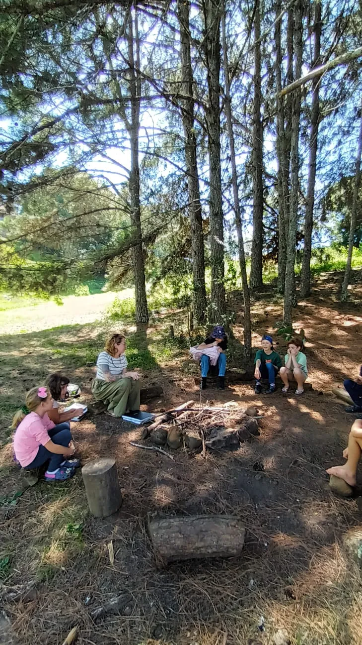 Roda de conversa com educadora e crianças ao redor de uma fogueira no bosque