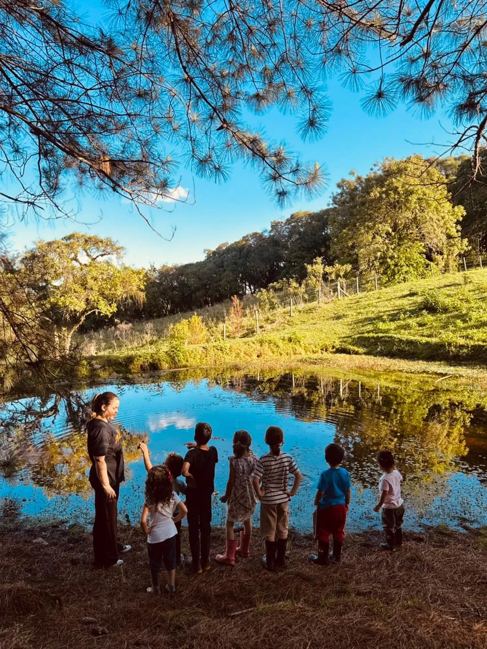 Educadora e um grupo de crianças observando as águas de um pequeno lago rodeado por natureza