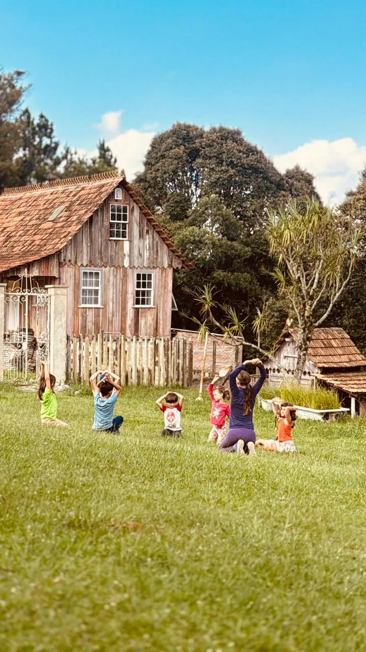 Educadora e um grupo de crianças fazendo alongamento na grama em frente a uma casa rústica de madeira